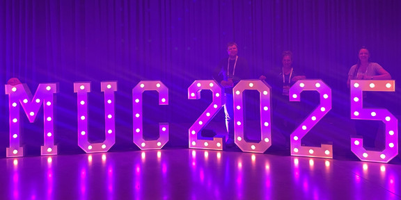 Violet illuminated letters stand out against a purple background. Behind them are Hendrik Sellmann, Frauke Mörike, and Jana-Sophie Effert leaning against the illuminated letters.