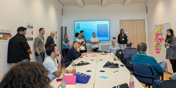 Eine kollaborative Arbeitssession mit vielen Teilnehmenden, bei welcher die Mehrheit an einem Tisch sitzt und eine Person ein Bulletin-Board mit Notizblättern vorstellt.