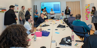 Eine kollaborative Arbeitssession mit vielen Teilnehmenden, bei welcher die Mehrheit an einem Tisch sitzt und eine Person ein Bulletin-Board mit Notizblättern vorstellt.