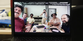 Zu sehen ist ein Laptop, welcher ein Bild von einem Plenum von Personen zeigt, die um einen Tisch herum sitzen und in die Kamera lächeln. In der Mitte steht Ronda Ringfort-Felner und nimmt mit der Kamera das Bild vom Laptop auf.