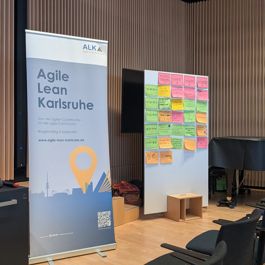 Ein großes Schild mit dem Text "Agile Lean Karlsruhe", daneben ein Bulletin-Board mit dem Tagesplan, unlesbar.
