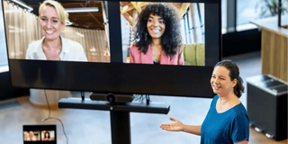 Eine Person, welche präsent im Raum steht, stellt zwei weitere Personen vor, welche auf einem Bildschirm zu sehen sind.