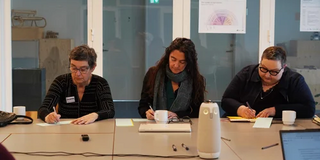 Drei Teilnehmende des Symposiums sitzen an einem Konferenztisch und schreiben.