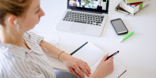 Am Schreibtisch sitzende Studentin schaut ein Lernvideo, daneben liegen Bücher und Stifte.